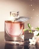 Frasco de perfume feminino elegante rosa com flores brancas e folhas ao lado, fundo iluminado.